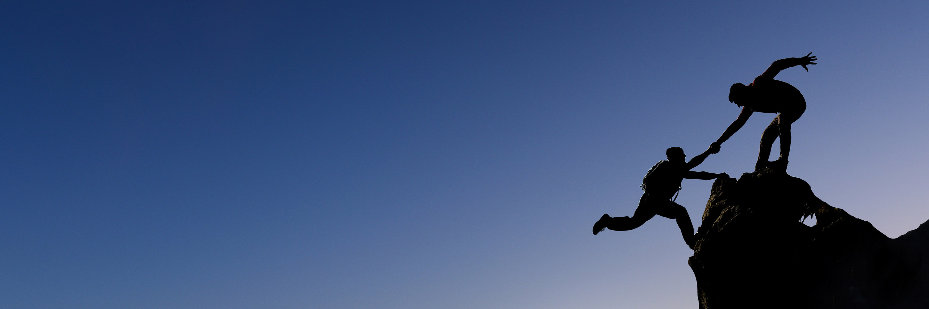 Hiker helping another hiker up a cliff in front of a twilight sky.