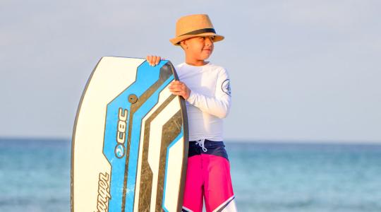Boy wearing hat with a surf board by the ocean.,